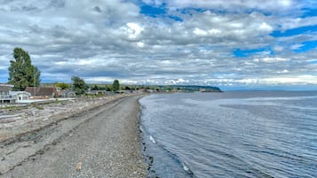 Una carretera costera con casas a la izquierda, que conduce a una playa con aguas tranquilas y un cielo nublado.