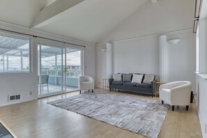 A spacious living room with a large sliding glass door leading to a balcony, a grey sofa with cushions, a white armchair, and a patterned rug. - Serene 3BR Waterfront Home Balcony With Bay Views (Point Roberts)