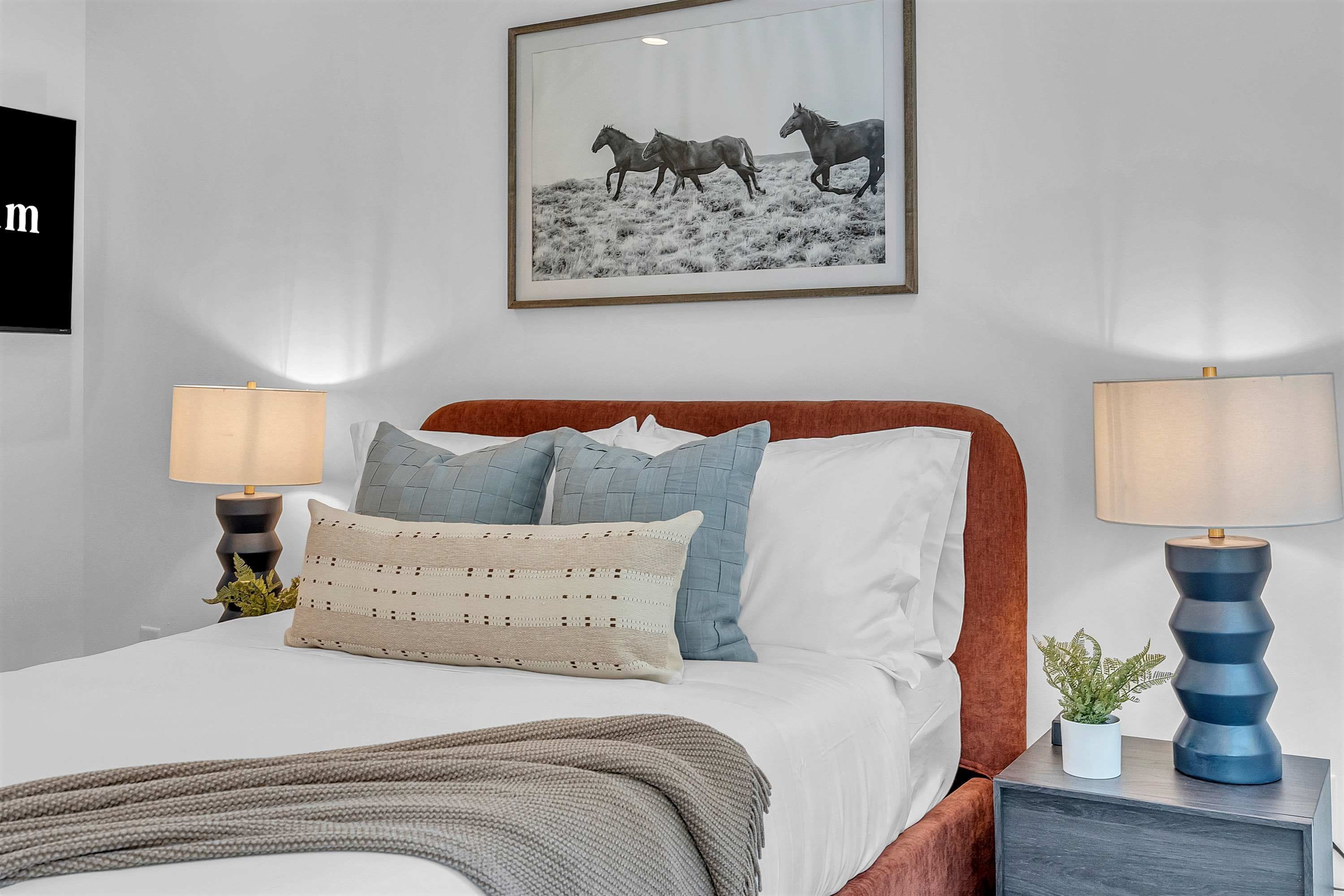 Cama bem arrumada com cabeceira marrom, duas mesas de cabeceira com abajures, um quadro na parede e uma pequena planta em uma das mesas.