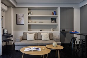Ein modernes Wohnzimmer mit einem grauen Sofa, zwei runden Holztischen, einem Bücherregal mit Dekoration und einer kleinen Küchenzeile mit Hockern.