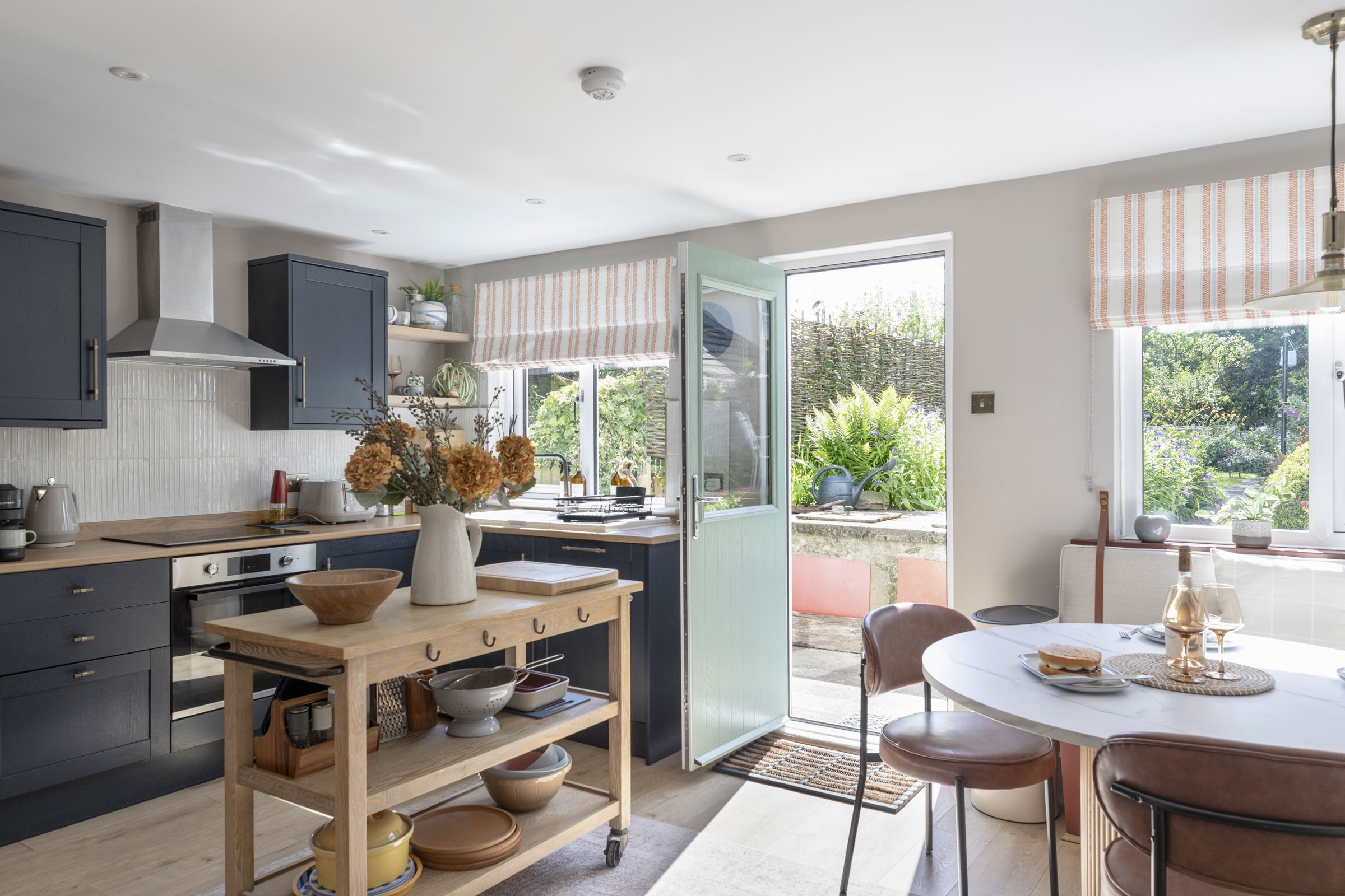 Een moderne keuken met een centraal eiland, een eethoek met een ronde tafel en uitzicht op de tuin door de open deur.