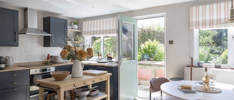 A modern kitchen with a central island, a dining area with a round table, and a view of the garden through the open door.