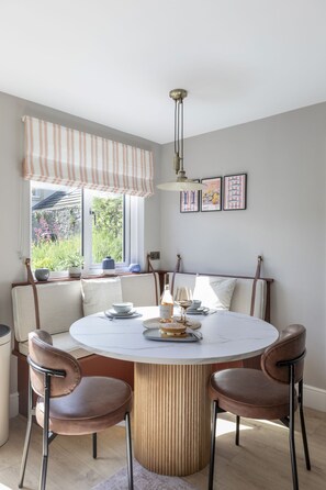 A round dining table with a white marble top, surrounded by four brown leather chairs with metal legs. Above the table, there is a pendant light fixture. The room has a window with a striped valance, and a view of greenery outside. - Orca Cottage, Seahouses, Northumberland (Seahouses)