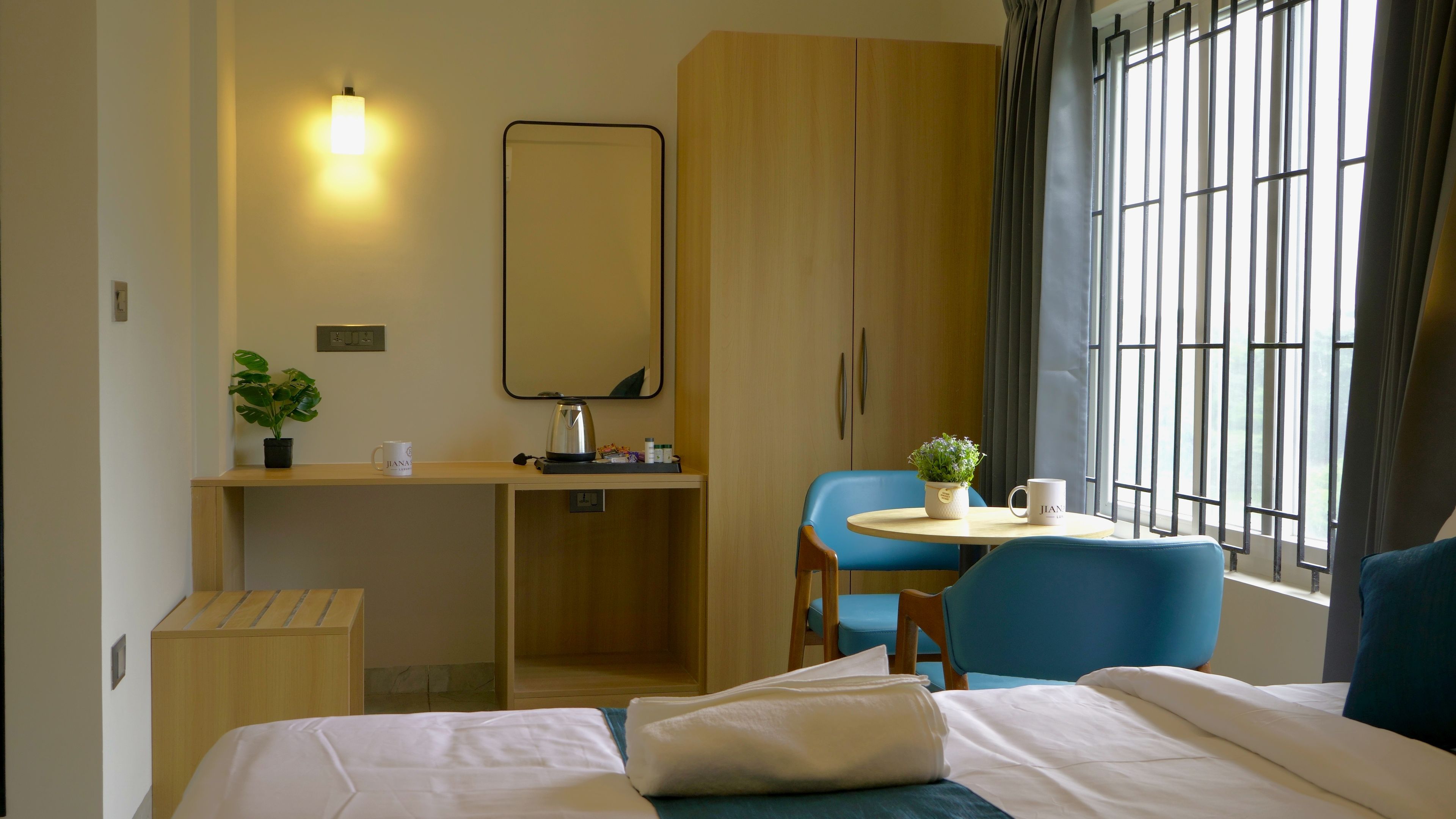 A hotel room with a bed, a desk, a chair, a small table with a plant, and a window with curtains.