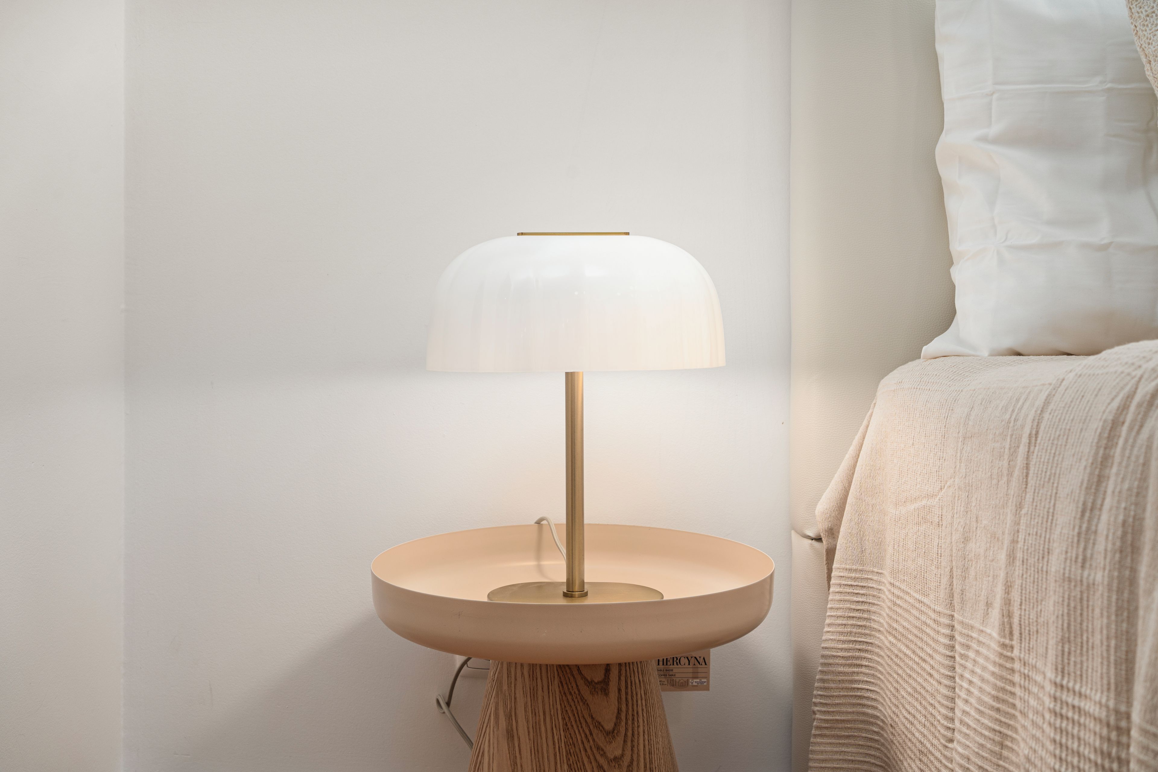 Une table de chevet avec une lampe, un lit recouvert d’une couverture blanche et un mur orné d’une étiquette.