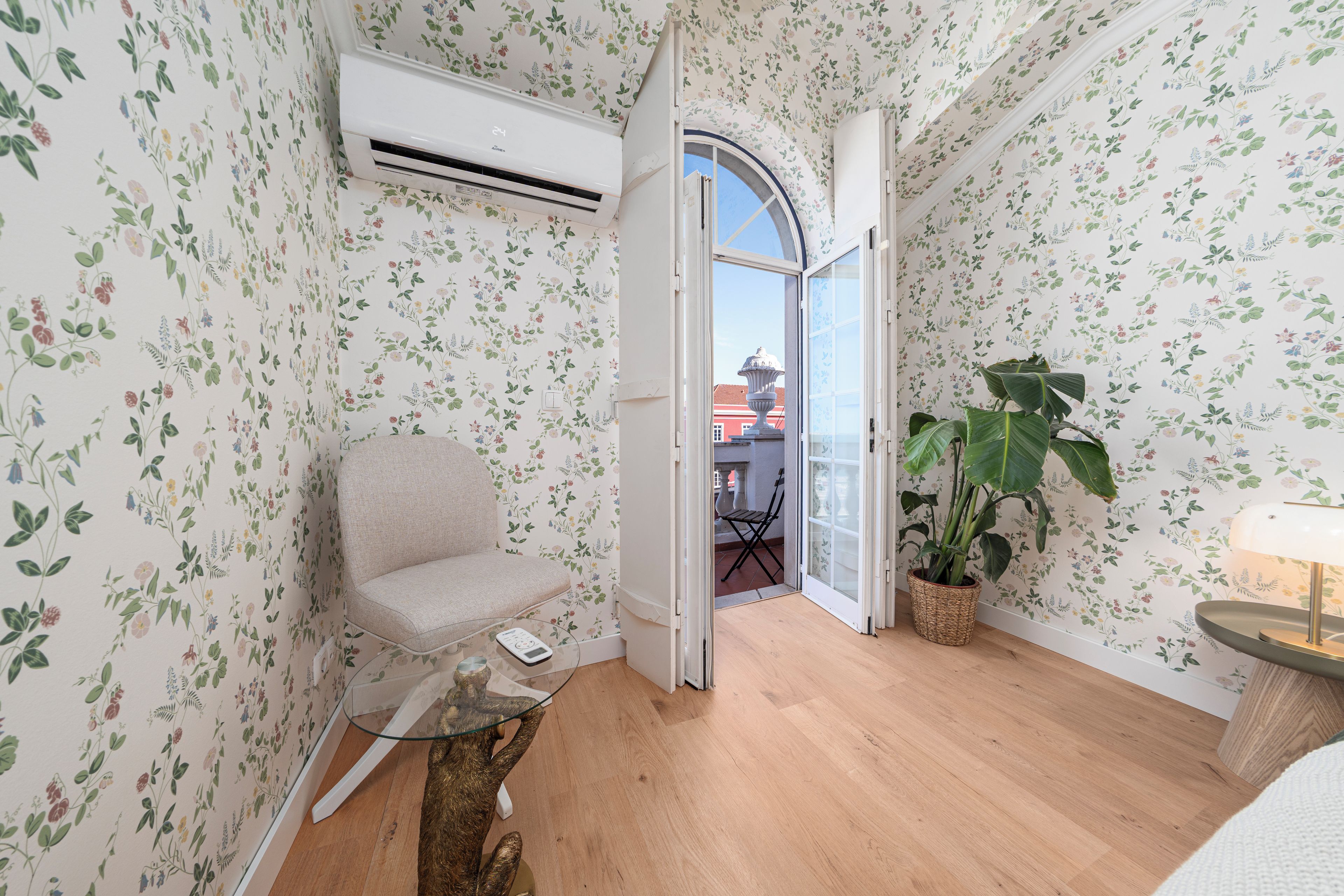 Une pièce avec un papier peint à motifs floraux, un plancher en bois, une chaise, une table avec une lampe et une plante en pot.