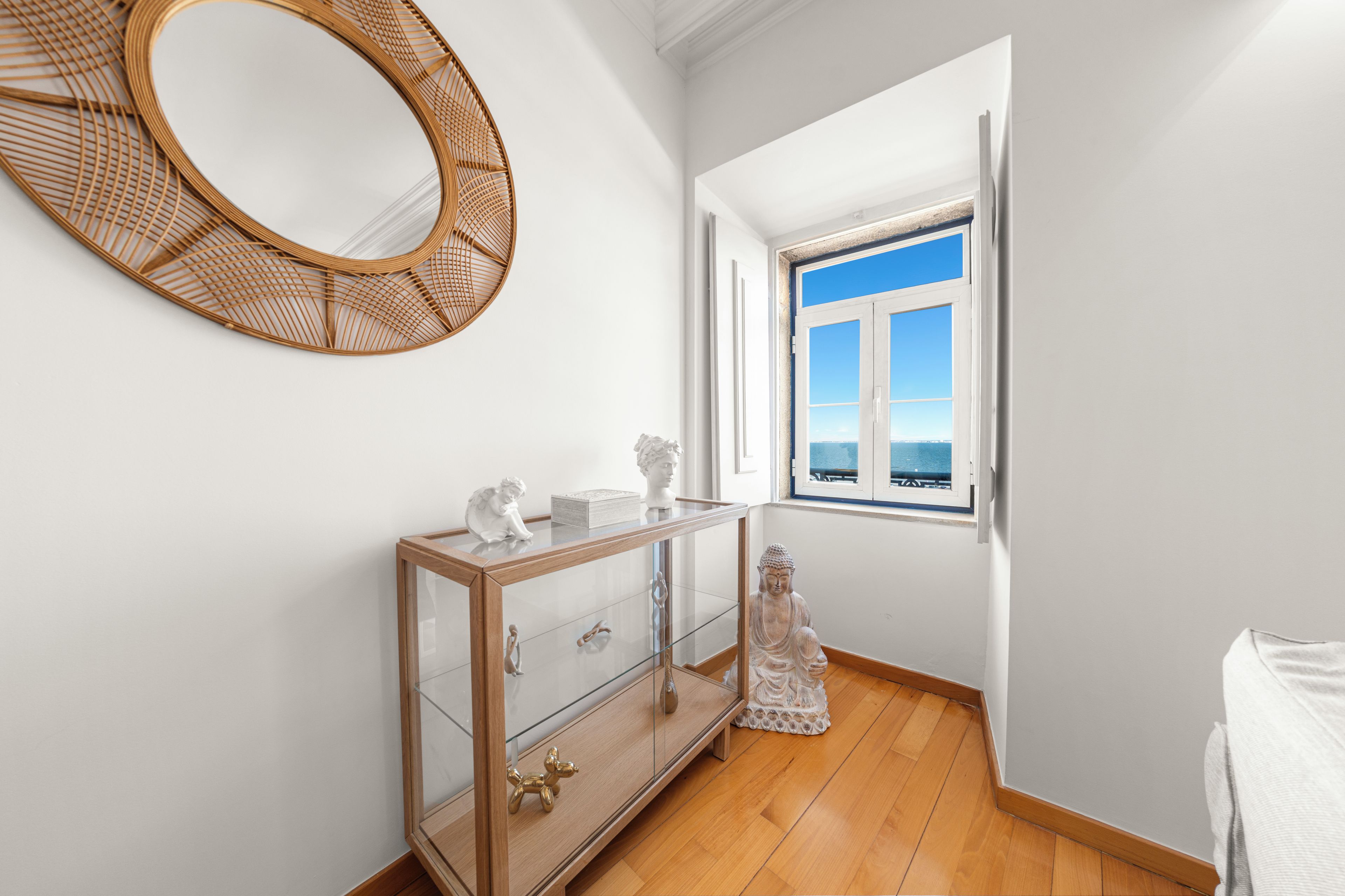 A room with a wooden floor, a glass cabinet with drawers, a decorative mirror, and a window with a view of the sea.