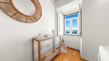 A room with a wooden floor, a glass cabinet with drawers, a decorative mirror, and a window with a view of the sea.