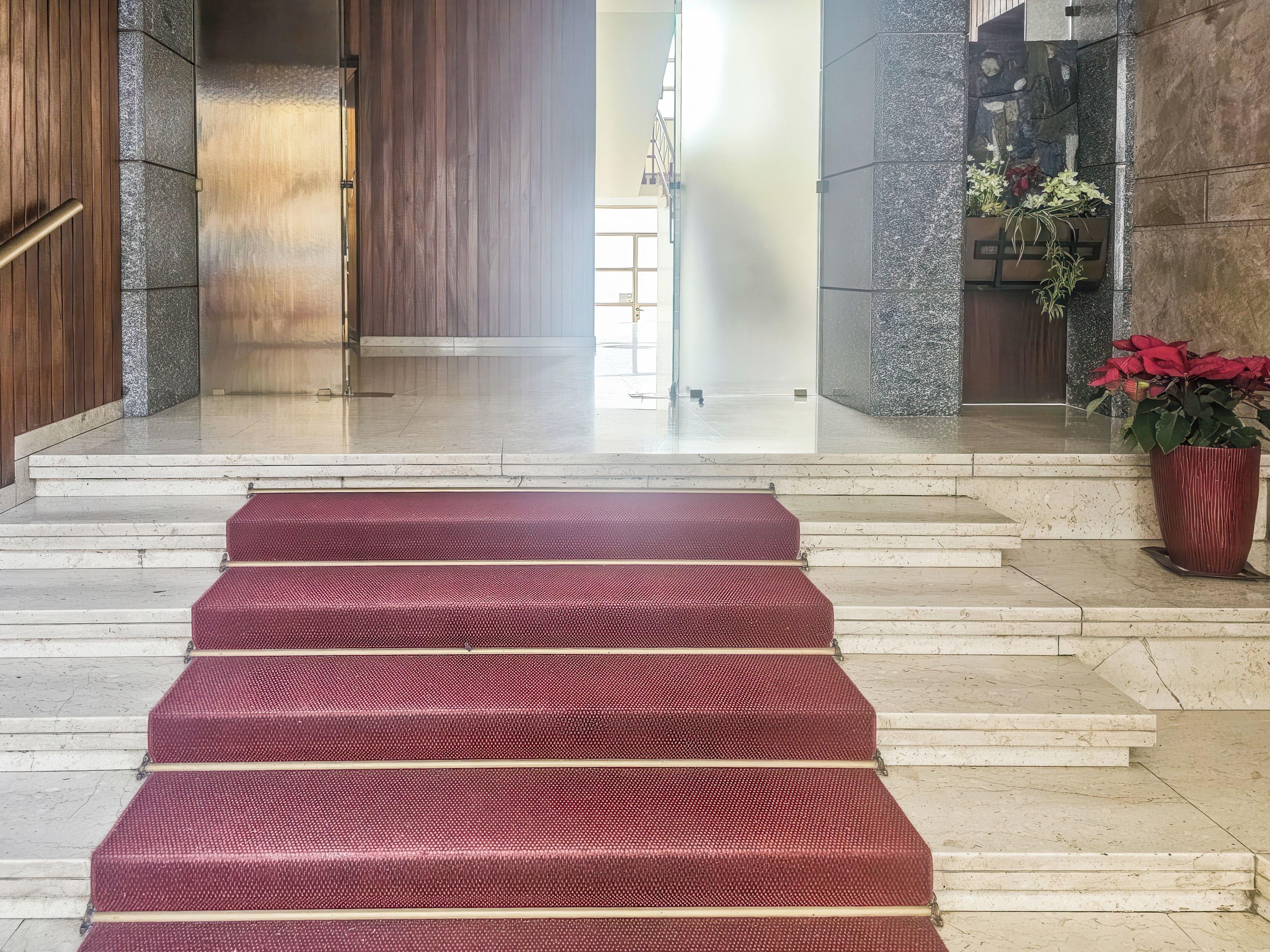 Unos escalones de mármol conducen a una puerta de madera, con un rellano alfombrado de rojo y una planta en maceta a la derecha.