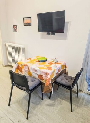 A dining area with a table covered in a floral tablecloth, two chairs, a television mounted on the wall, and a radiator. - AL NIDO (Torino)