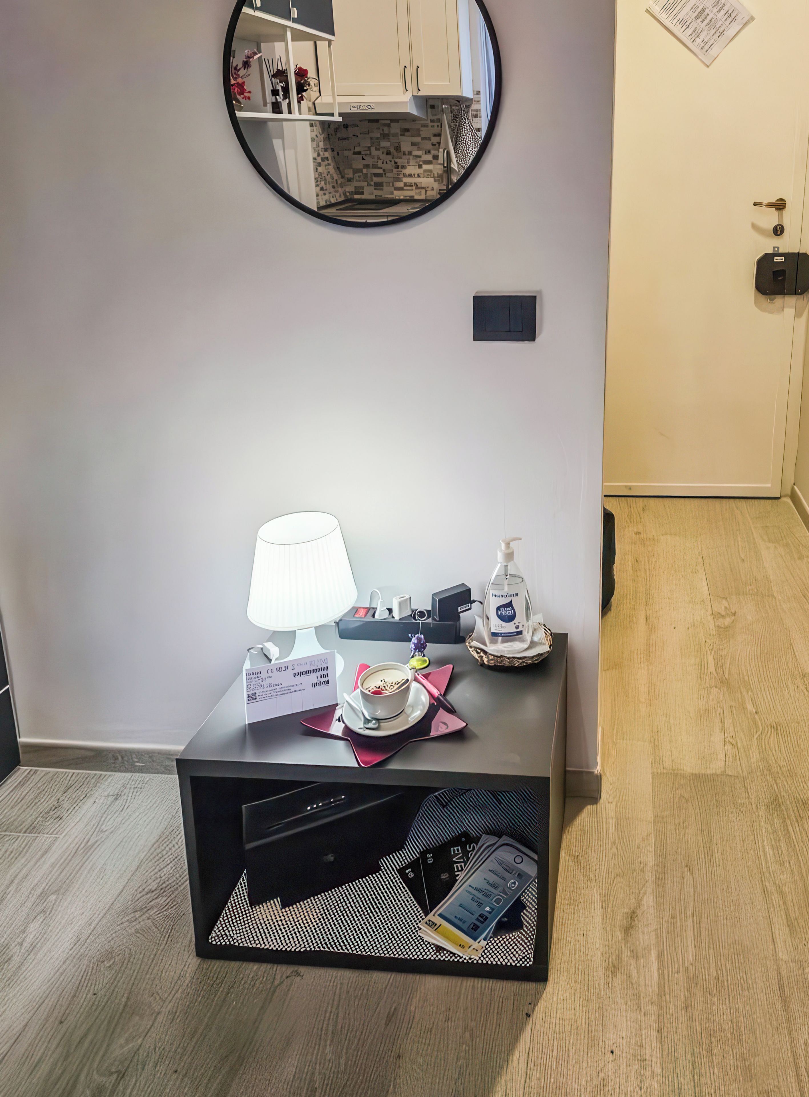 Una habitación con un espejo redondo, una mesita con una lámpara, una taza de café, una botella de jabón de manos y una cesta con pañuelos.