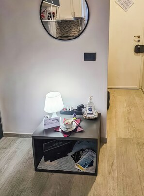 A room with a round mirror, a small table with a lamp, a cup of coffee, a bottle of hand soap, and a basket with a tissue. - AL NIDO (Torino)