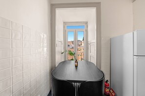 Eine schmale Küche mit weißen Fliesen, einem schwarzen Tisch und Blick durch eine Tür nach draußen.