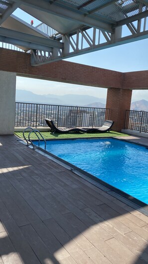 An outdoor swimming pool with a view of distant mountains, surrounded by a wooden deck and metal railings. - Cozy 1-bedroom apartment in beautiful Santiago with AC (Santiago)