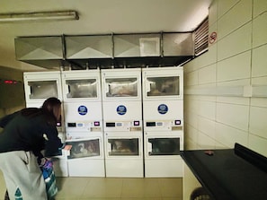 A laundromat with multiple washing machines and dryers, a person using one of the machines, and a no-smoking sign on the wall. - Cozy 1-bedroom apartment in beautiful Santiago with AC (Santiago)