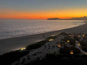 Paisatge costaner amb una platja, un camí i un petit assentament amb edificis il·luminats.