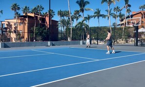 Een tennisbaan buiten met twee spelers, omgeven door palmbomen en gebouwen.