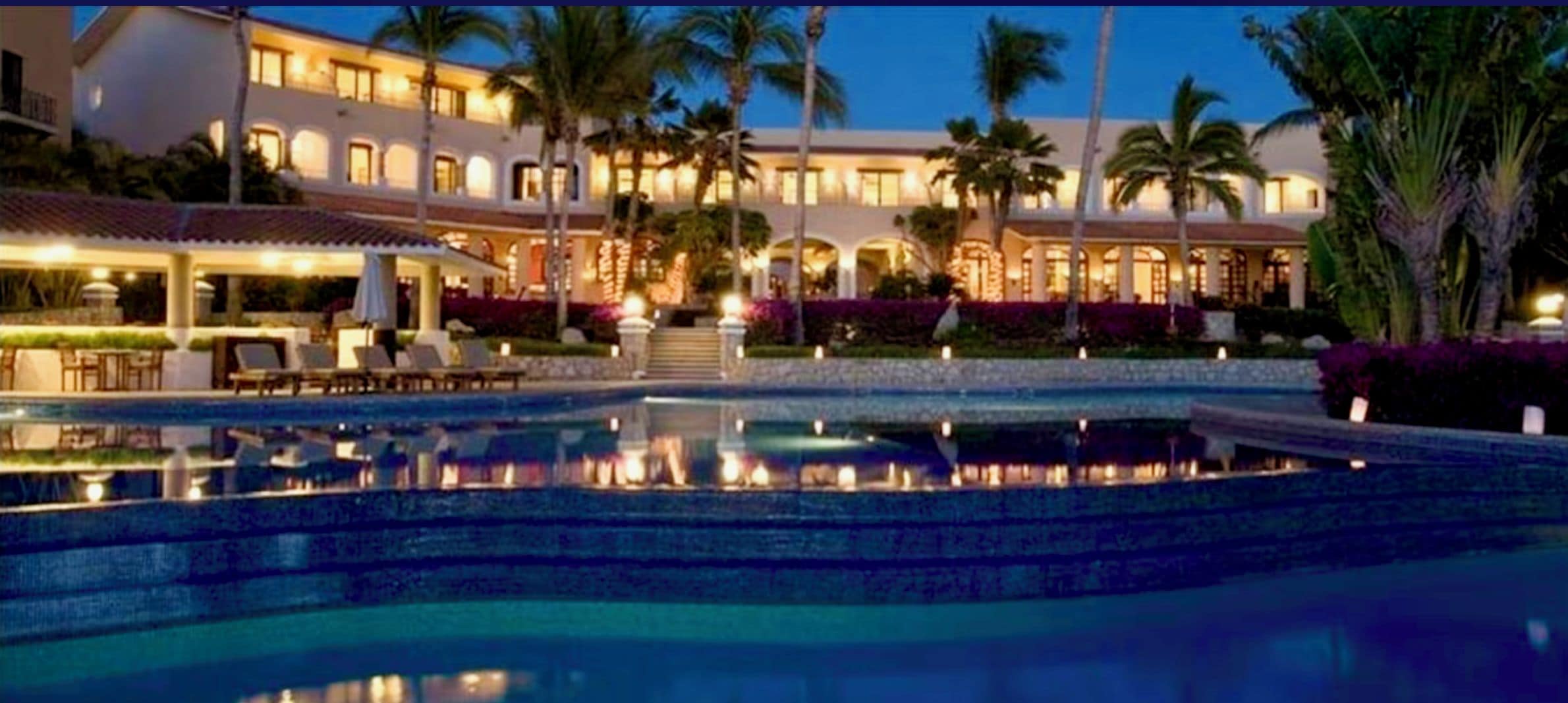 Un hôtel doté d’une piscine, de palmiers et d’une façade bien éclairée.