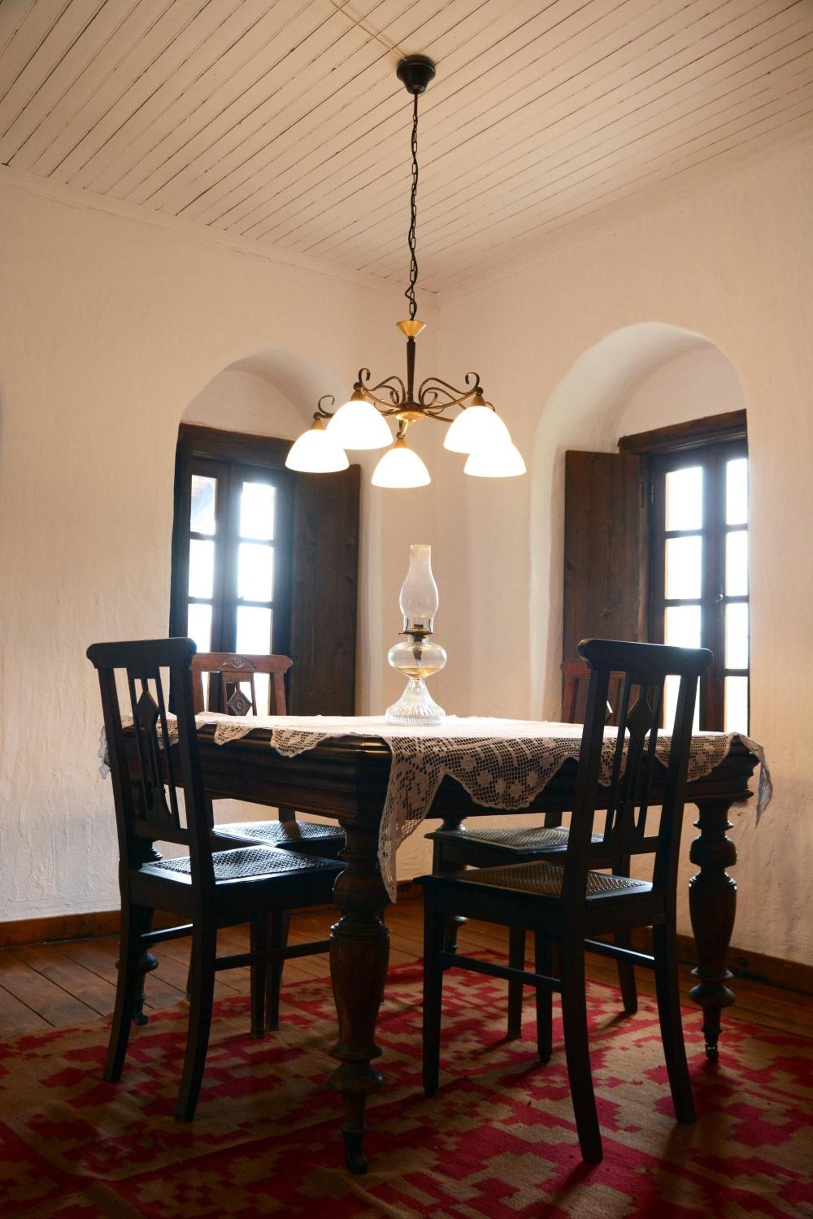 Une salle à manger avec une table dressée pour quatre personnes, un lustre et deux fenêtres avec des rideaux.