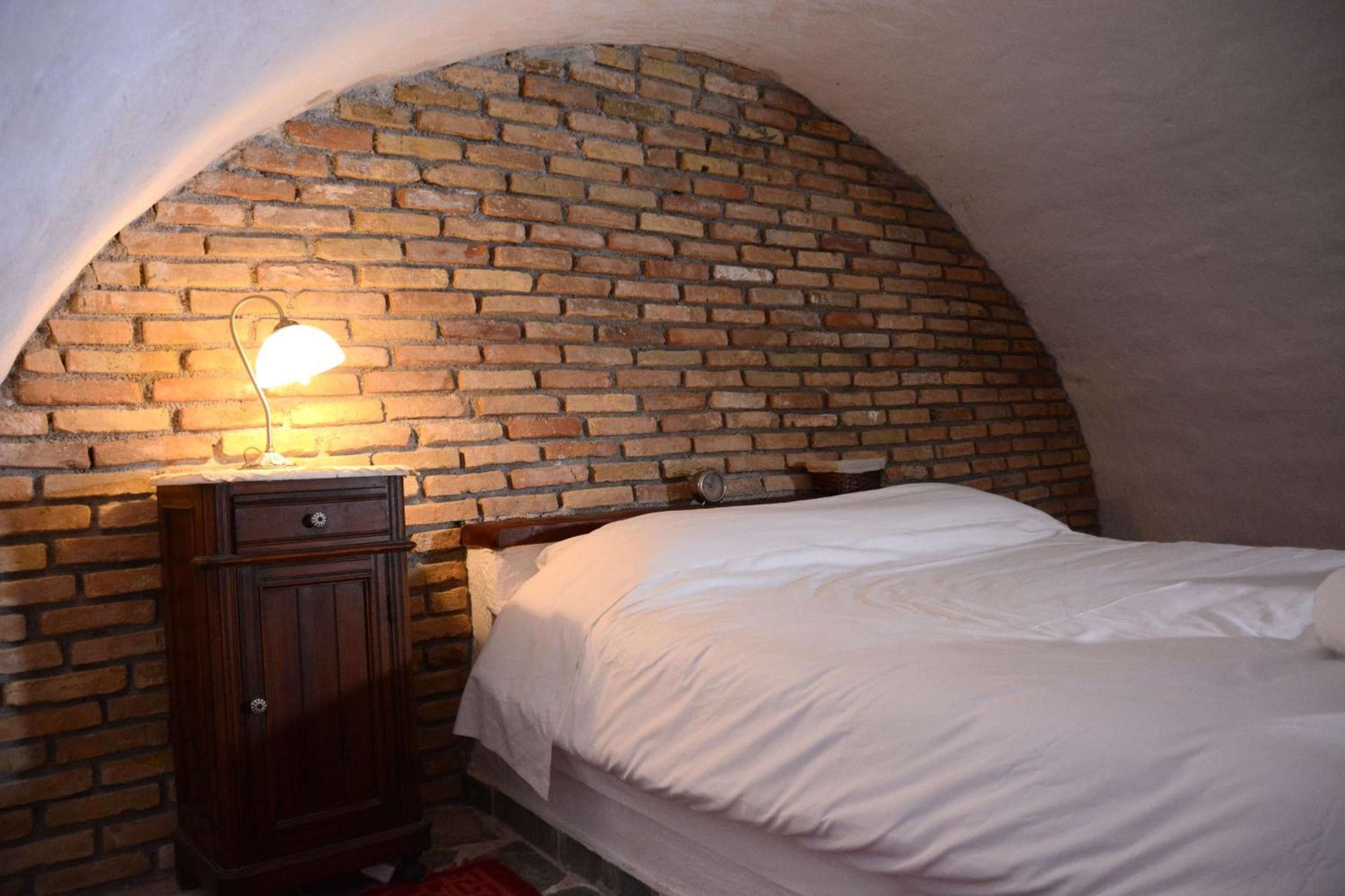 A bedroom with a brick arched ceiling, a wooden nightstand with a lamp, and a bed with white bedding.