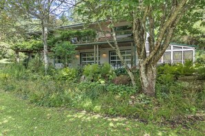 A house with a porch, surrounded by dense vegetation and trees. - Stunning 3,000 sq ft Blue Ridge Pkwy Guest House 30 Acres/Pond/Hot Tub/Game Room (Floyd)