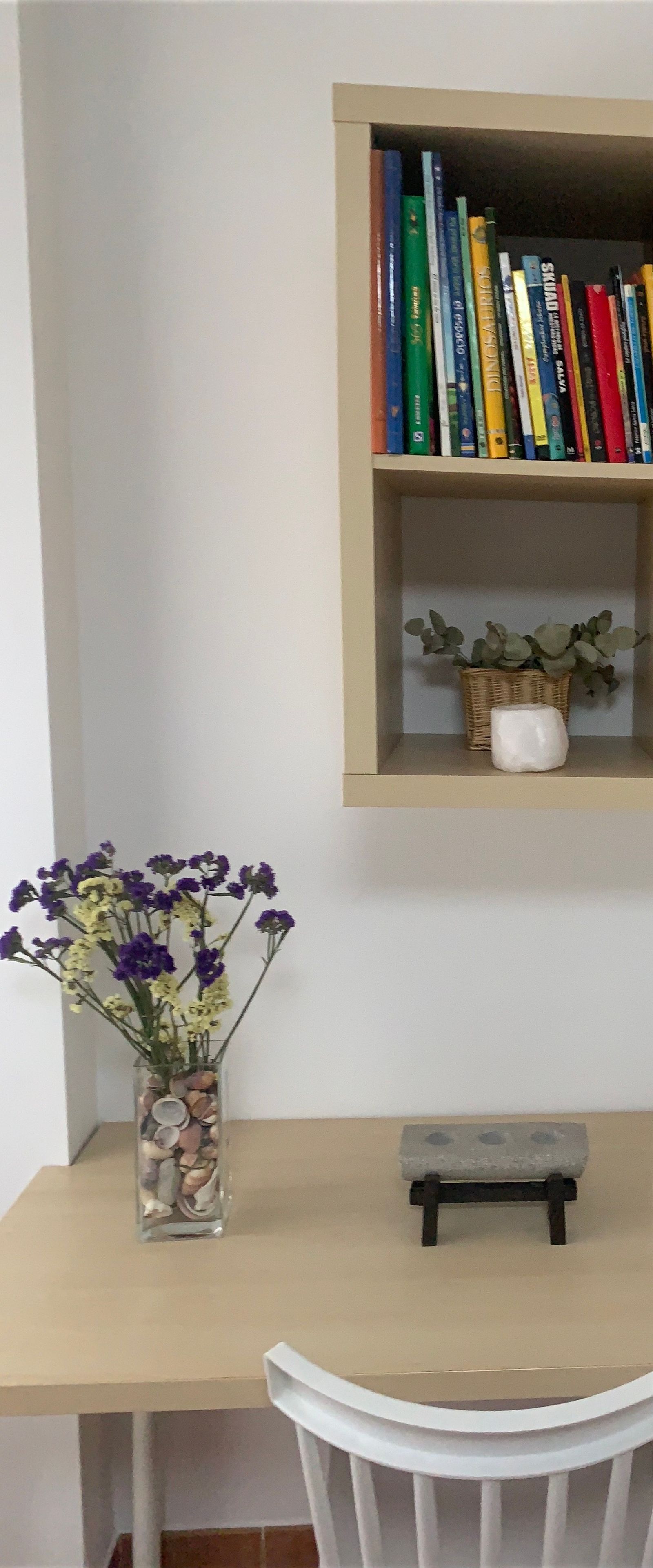 Una habitación con un escritorio, una silla, una estantería y un jarrón con flores.