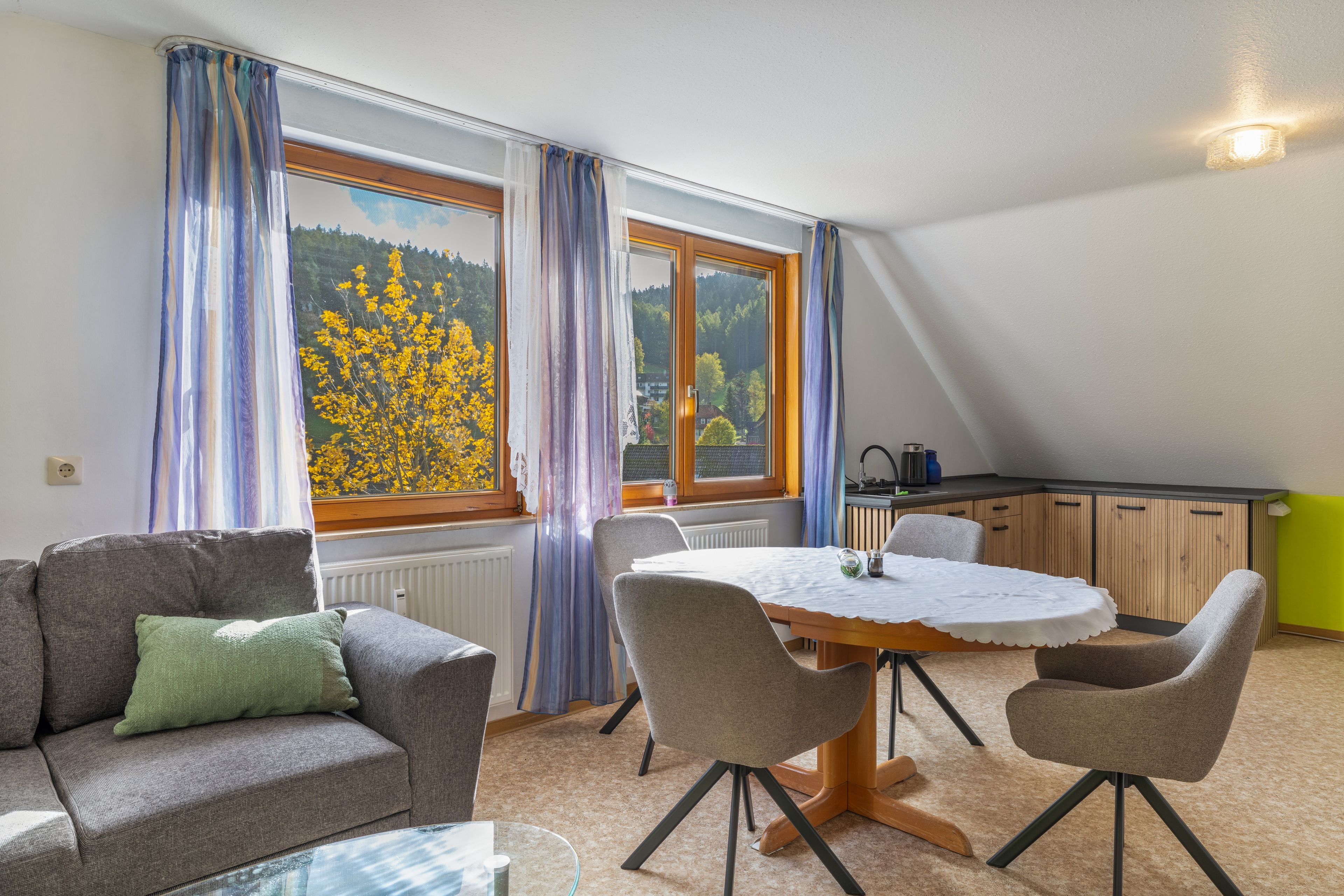 Ein gemütliches Zimmer mit Esstisch, Stühlen, einer Couch und Blick auf einen Wald durch große Fenster.