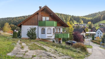 Una casa de dos pisos con techo inclinado, un balcón y una pequeña construcción adosada con techo verde.
