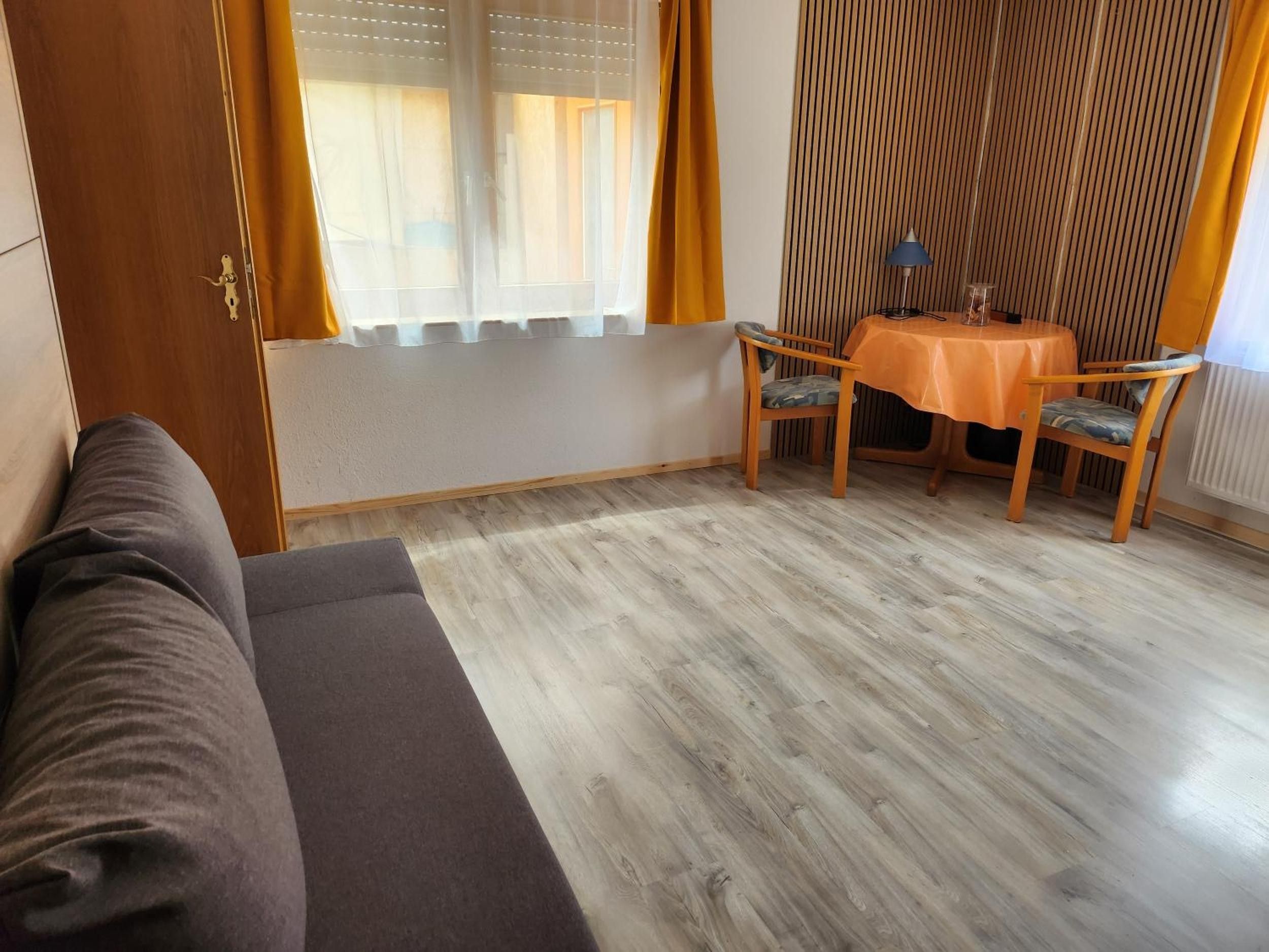 Een kamer met een bank, een tafel met stoelen, een raam met gordijnen en een houten wandpaneel.