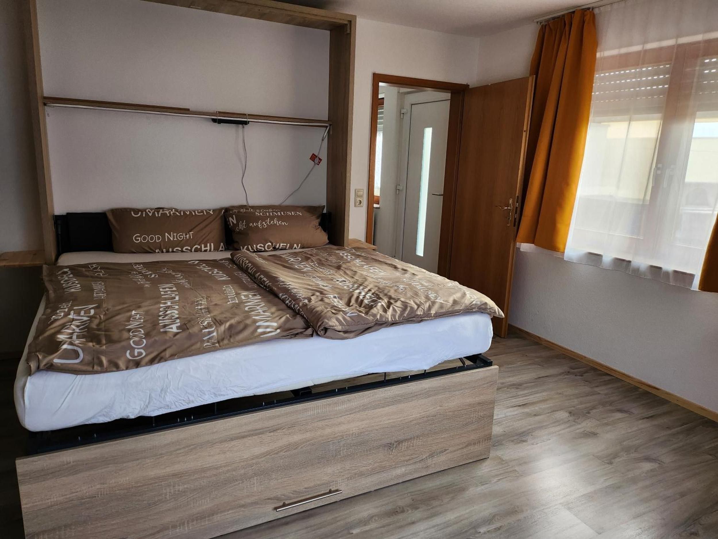 A bed with a wooden frame and a storage drawer, a bedside table, and a window with curtains.