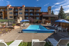 A pool area with lounge chairs and umbrellas, surrounded by a multi-story building with balconies. - Slopeside | Washer/Dryer | Hot Tub & Pool | On Bus Route (Steamboat Springs)