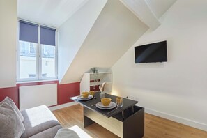 A modern living room with a dining area, a TV mounted on the wall, and a window with blinds. - Charming Studio - Center of Paris Montorgueil (Paris)