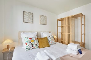 A hotel room with a bed, pillows, a bedside table with a lamp, a wooden wardrobe, and a stack of towels. - Charming apartment - Center of Paris Montorgueil (Paris)