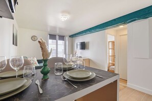 A dining area with a table set for four, a clock on the wall, and a television mounted on the wall. - Charming apartment - Center of Paris Montorgueil (Paris)