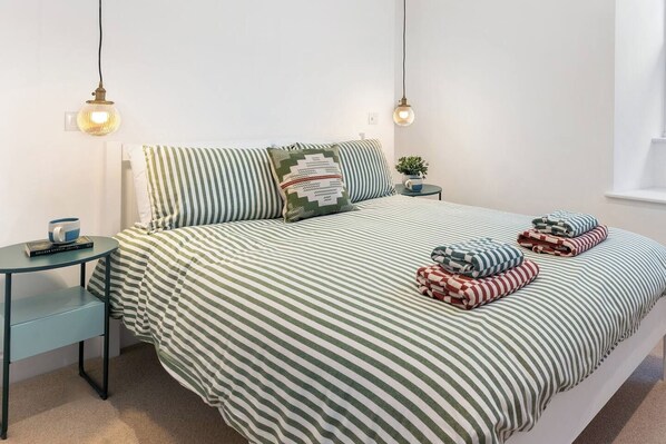 Een net opgemaakt bed met gestreepte lakens, een nachtkastje met een mok en boek, en twee hanglampen.