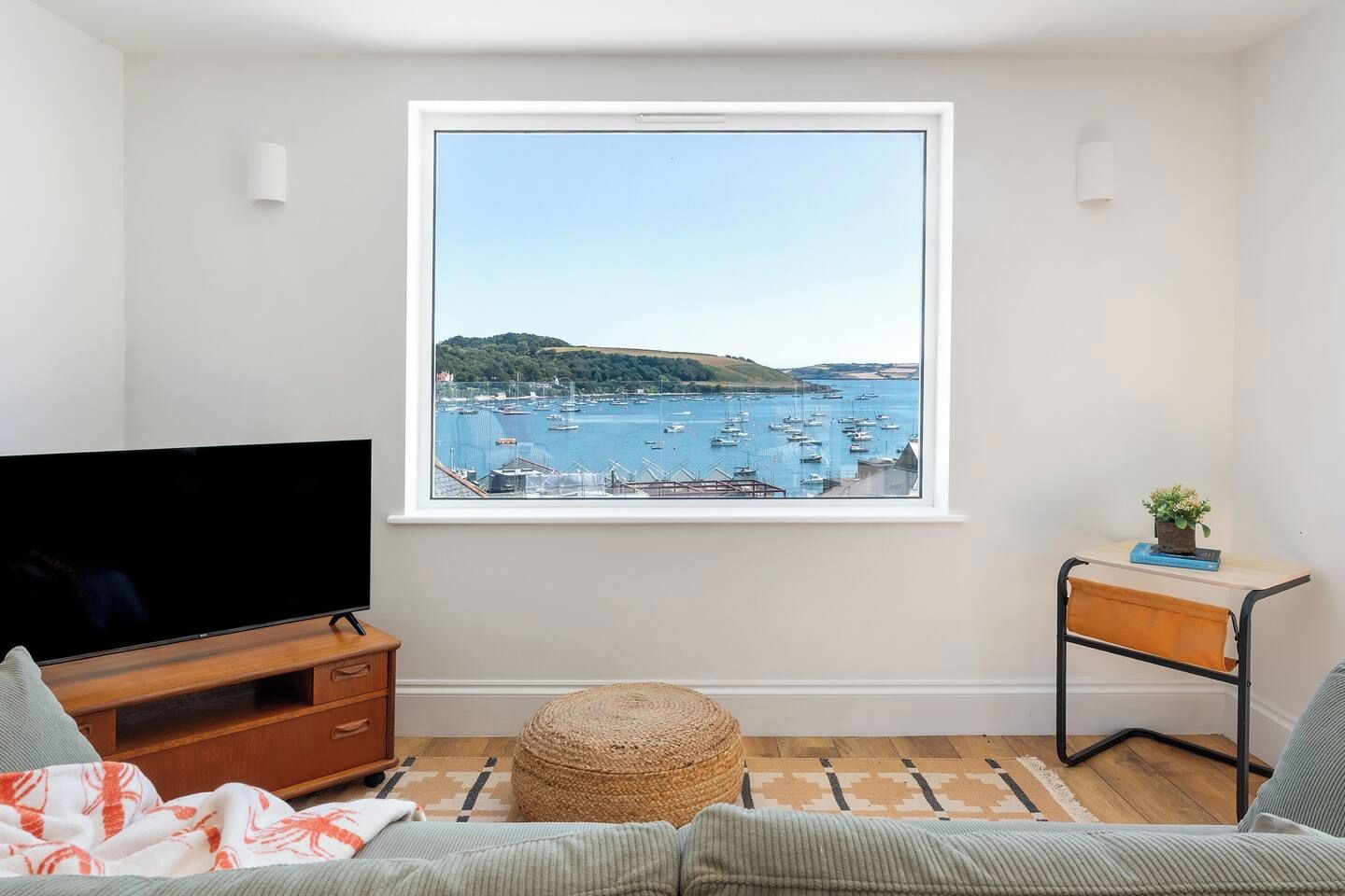 Un salon comprenant un téléviseur, un canapé, une petite table avec une plante, et donnant sur un port où sont amarrés des bateaux.