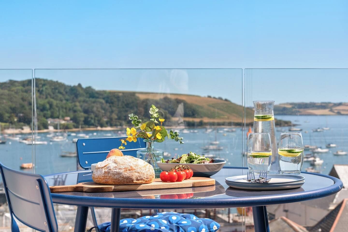 A table with a loaf of bread, a bowl of salad, a vase with flowers, and glasses of water, set against a scenic harbor view.
