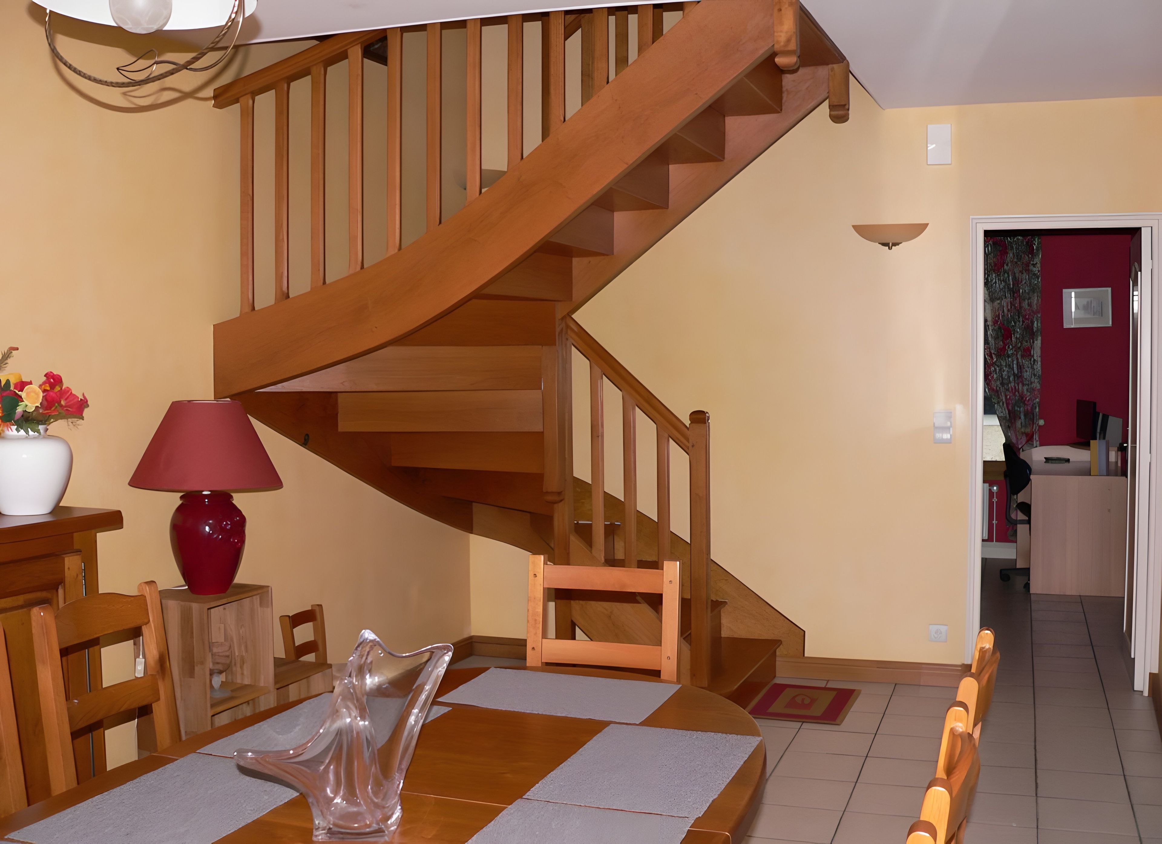 Un escalier en bois menant à l’étage supérieur, un coin repas avec une table et des chaises, et une lampe rouge posée sur une table d’appoint.