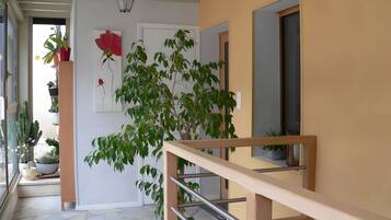 Un couloir avec un escalier, des plantes en pot, un tableau représentant une fleur rouge et une porte avec une fenêtre.