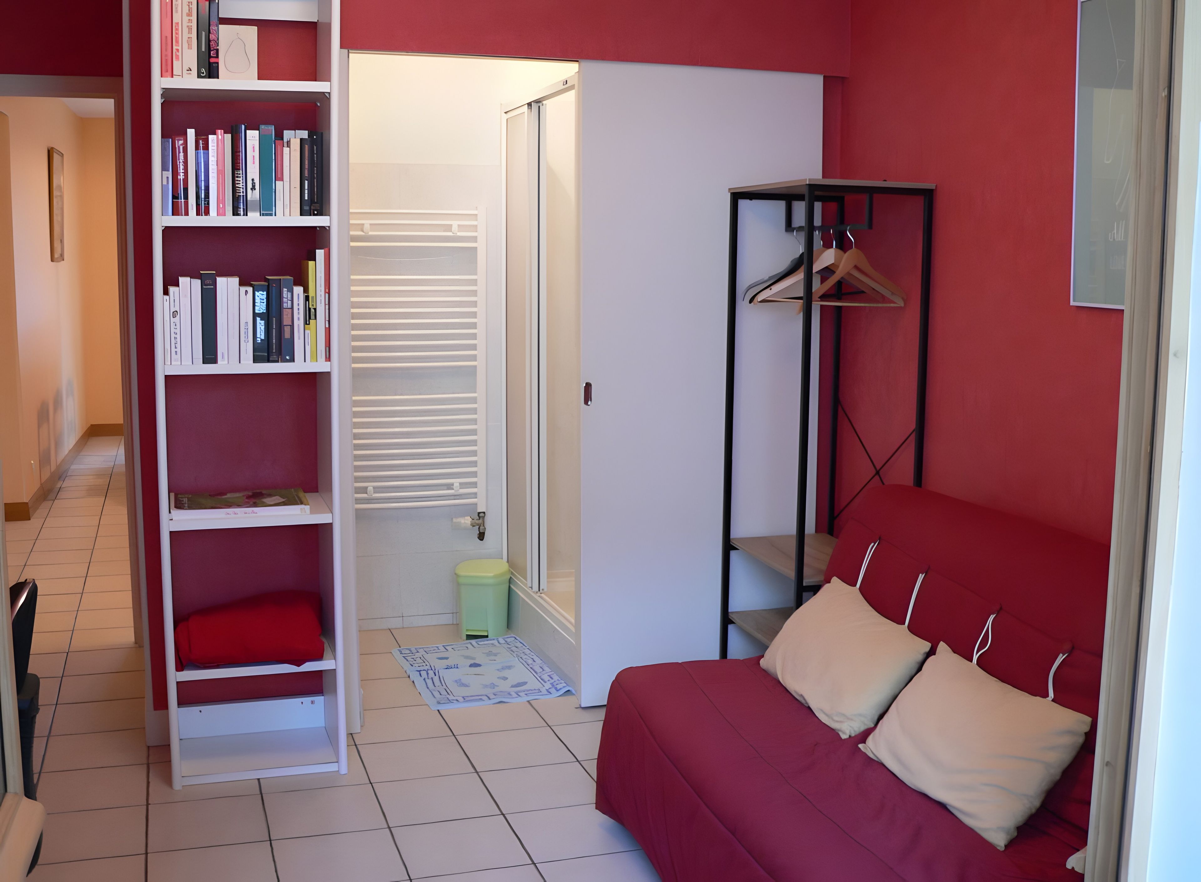 Une pièce avec un canapé rouge, une bibliothèque blanche, un porte-manteau noir et une porte qui donne sur une autre pièce.