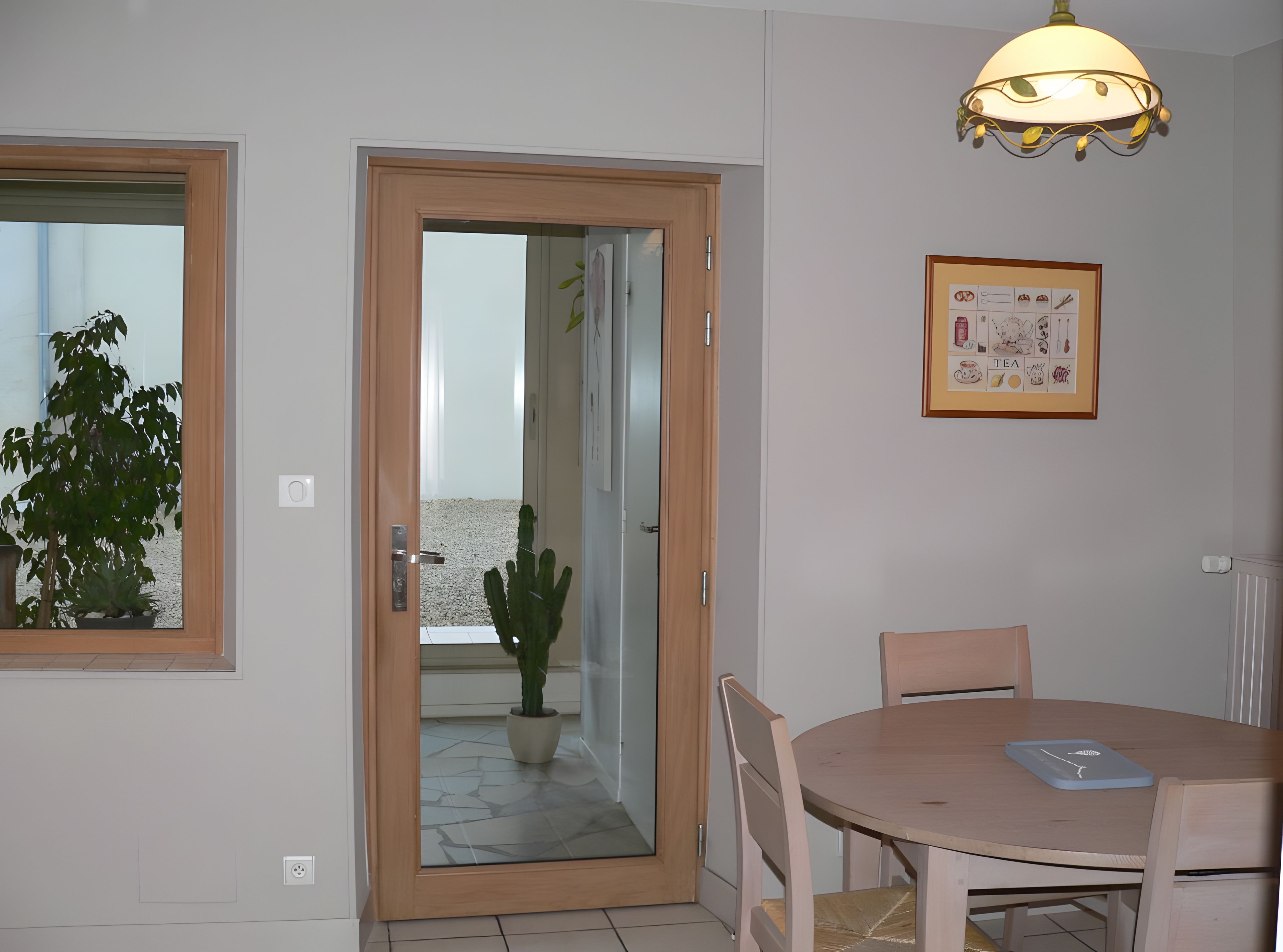Een kamer met een houten deur, een kamerplant, een lijst met een foto aan de muur en een ronde tafel met stoelen.