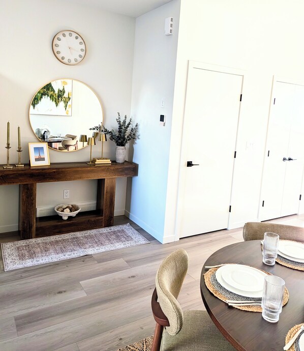 A dining area with a round table set for four, a wooden console table with decorative items, a large mirror, and a white door.