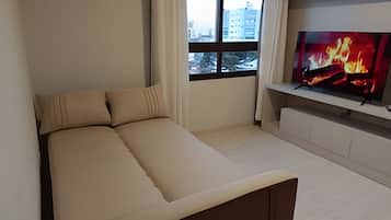 Un dormitorio moderno con una cama, un televisor sobre un mueble bajo y una ventana con vistas a edificios.