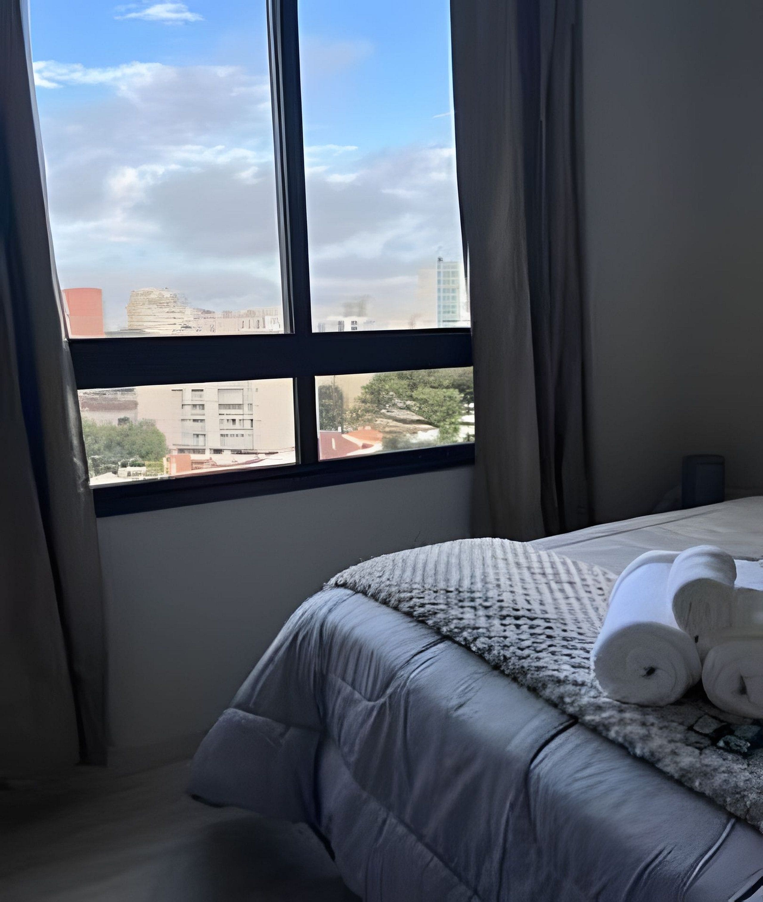 A hotel room with a bed, a window with curtains, and a view of buildings.