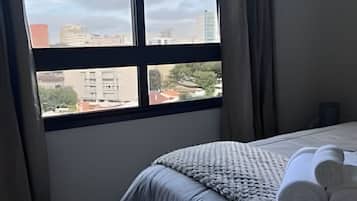 A hotel room with a bed, a window with curtains, and a view of buildings.
