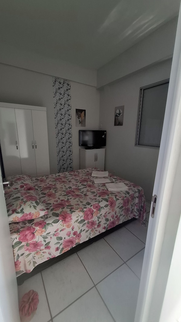 Un dormitorio con cama, armario, televisor sobre un mueble y una ventana.