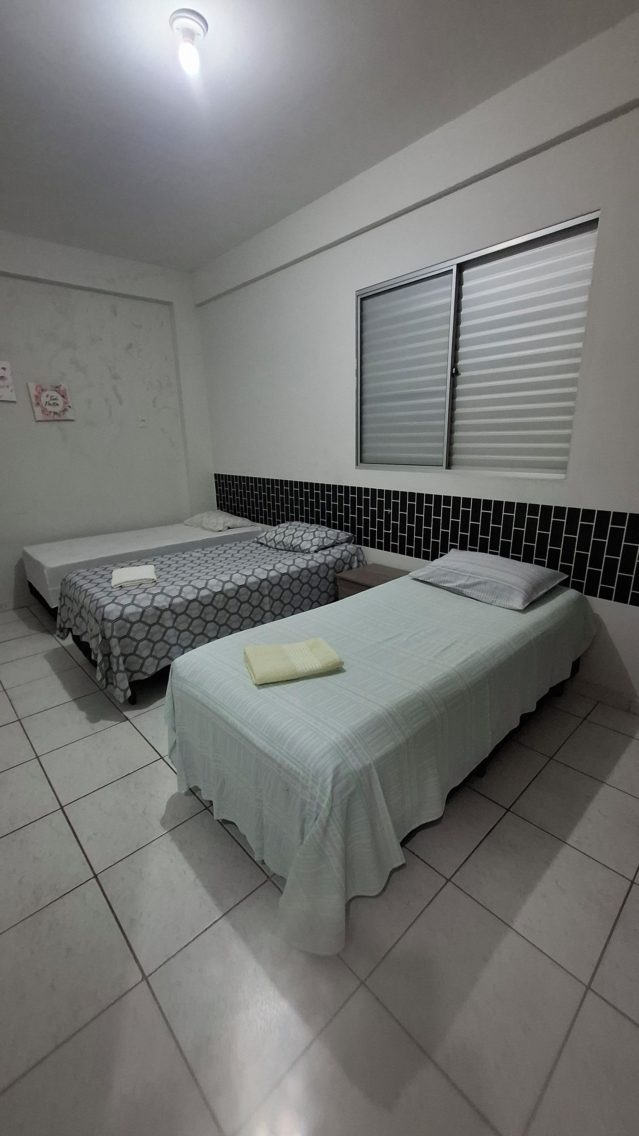 Una habitación con tres camas, una ventana con persianas, suelo de baldosas y una lámpara fijada a la pared.