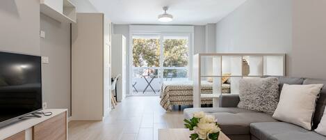 Una sala moderna con televisor de pantalla plana, un sofá gris, una mesa de centro de madera con un florero y un balcón con mesa y sillas.
