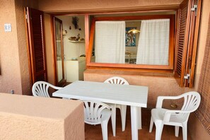 Une terrasse avec une table et des chaises blanches, une fenêtre avec des rideaux et une porte dotée d’un volet en bois.