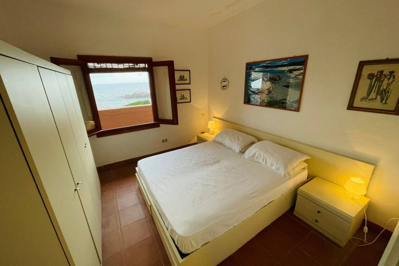 A bedroom with a bed, a nightstand, a window with a view of the sea, and a wardrobe.